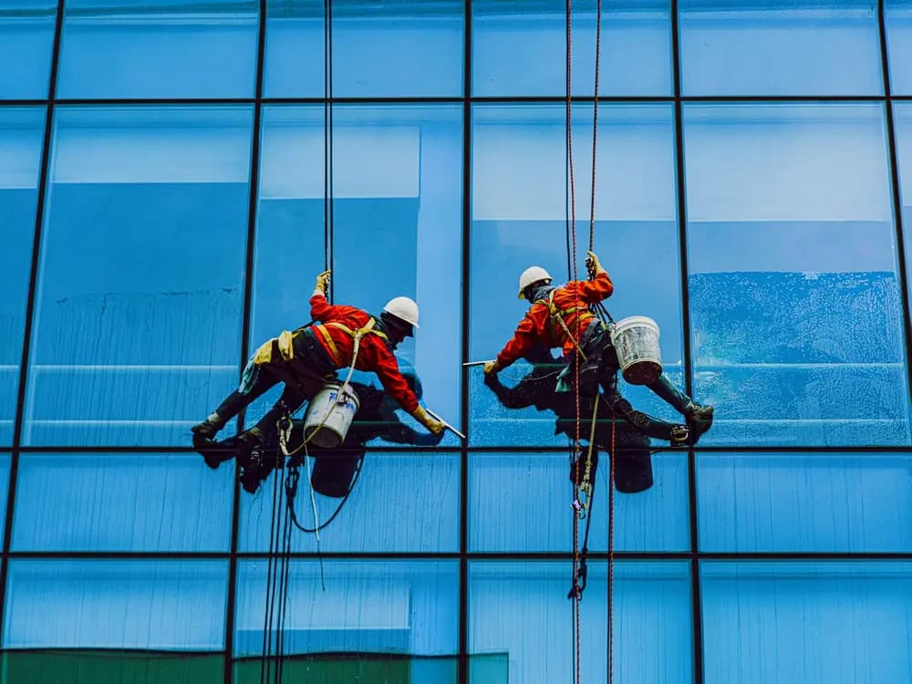 Dos técnicos limpiando la fachada de cristal de un edificio de oficinas suspendidos en cuerdas